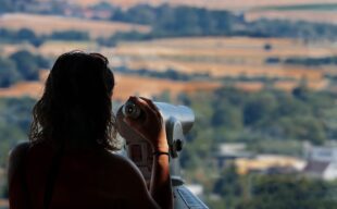 Silhouette of person looking through telescope to a leafy landscape of rolling hills.